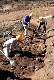 Lower part of the dip begins to take shape. After being armored with rock, workers begin to pull soil down over the rocks to a height water cannot go over it. Below, other workers fill in gully with more rock.
